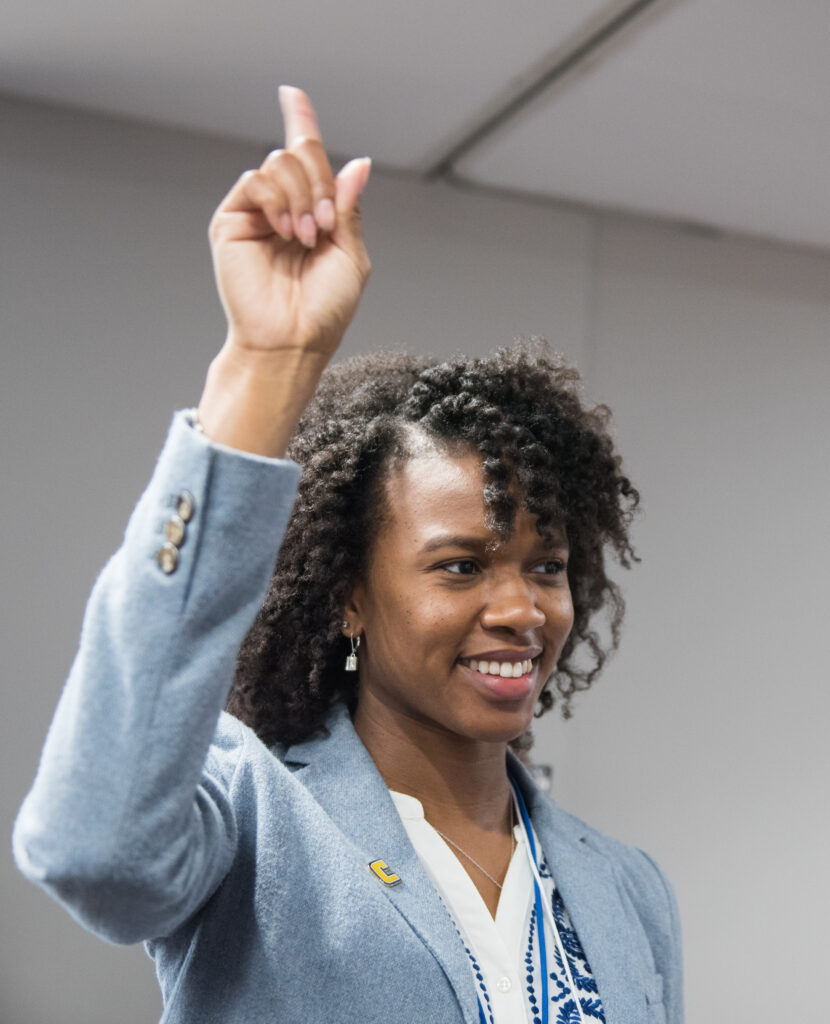Young woman in a light blue suit raising her hand. 