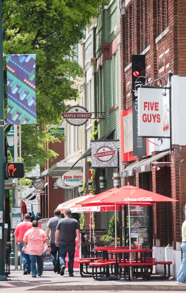 A group of four people walk down Broad Street in downtown Chattanooga, Tenn., surrounded by cafe tables; tall, red brick buildings; and a number of business signs.