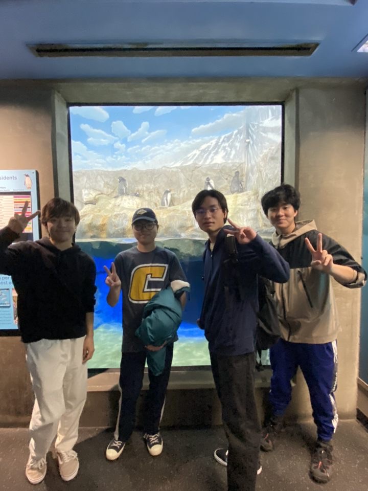 Four students pose in front of the penguin enclosure.