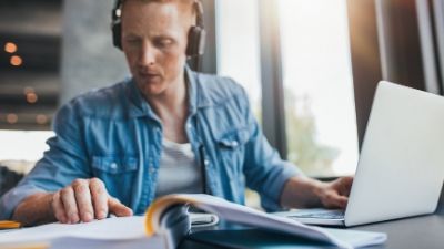 young man wearing headphones looking at a book with his laptop open