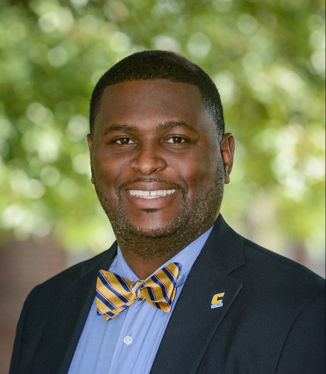 Bryan Samuel headshot. He is smiling, wearing a suit, with a very spiffy blue and gold bow tie.