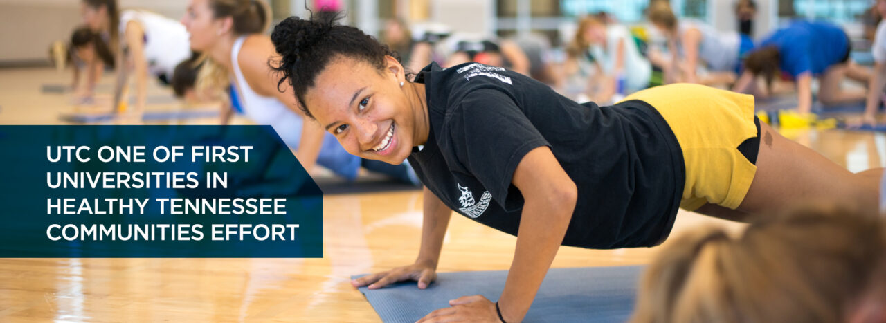 background image: UTC Student smiles for the camera while doing pushups in a Campus Recreation fitness class.