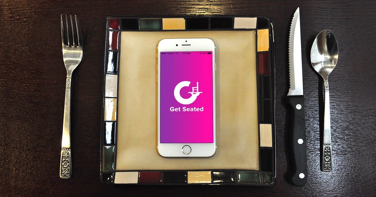 Photo of a smart phone displaying the Get Seated logo (a white G on a purple/pink background) laying on a plate with cutlery on either side.