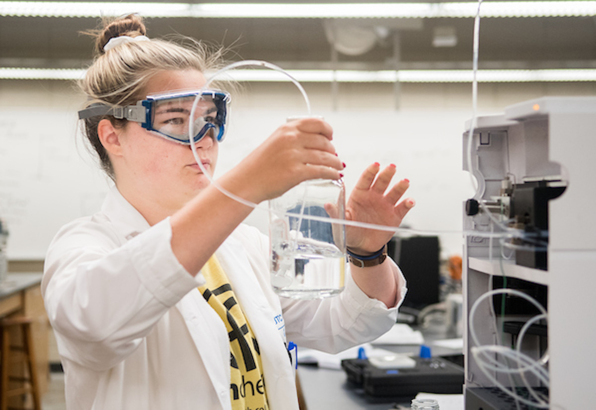 Student Samantha Sweck, in a lab coat and safety goggles holding a glass container with tubing.