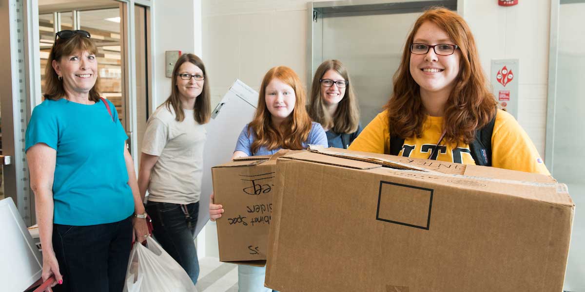 female freshman holds box and smiles as she moves into her dorm during operation move in, her family stands behind her carrying bags and boxes