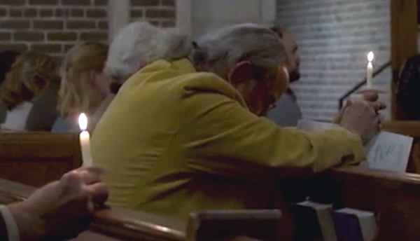 A man in yellow sits in a pew bowing his head and holding a candle.