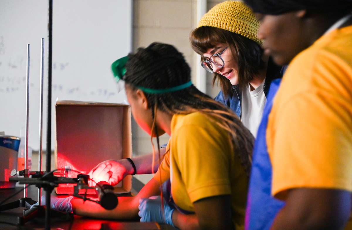 A UTC student holds her hand in front of a red lights and explains concept to two middle school students.