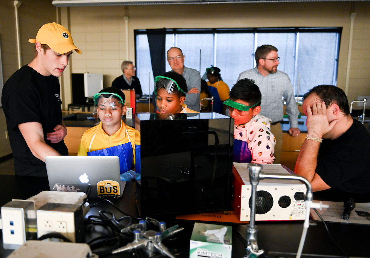 3 middle school students look at a laptop in a room full of technology. A UTC student shows them something on the computer.