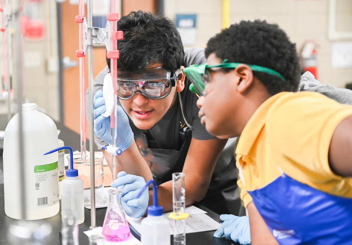 Two middle school students wearing safety goggles pour a pink liquid into a chemistry flask.