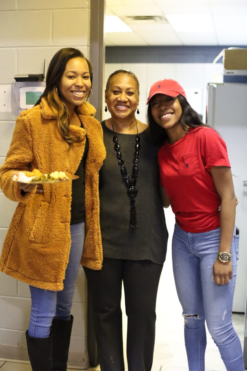 Shir Gholston smiles stands with her arms around two women.