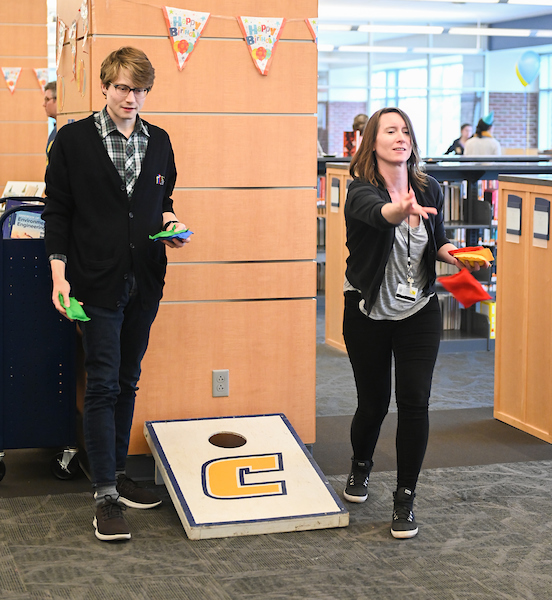 Two UTC students standing next to a cornhole board toss bags in the library.