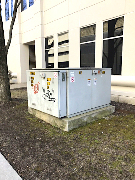 A large blank electrical box is on the ground next to a white building.