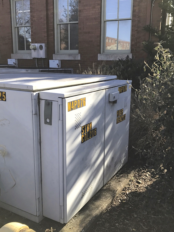 A large blank electrical box is on the ground near a large bush.