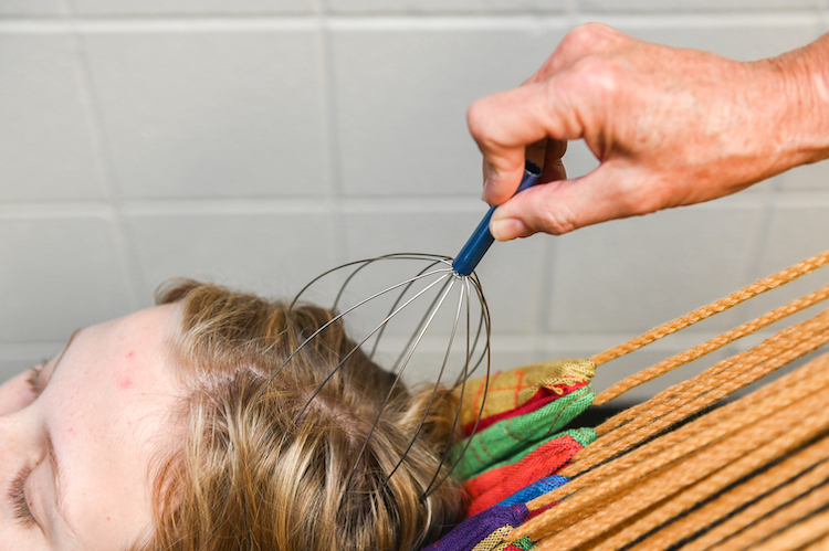 A student lays on a hammock while a hand uses a head scratcher on the laying student.