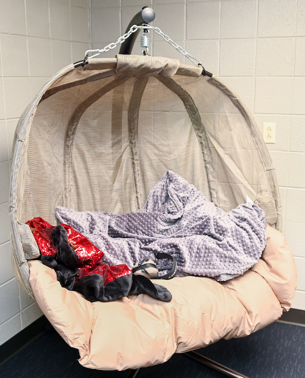 A large cream canopy chair is suspended from a metal pole in the corner of a room. The chair has a soft cushion and is covered in blankets.