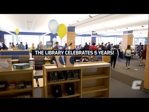 Text reading, "The Library Celebrates 5 years!" Many students stand in the library with balloons hung around the room