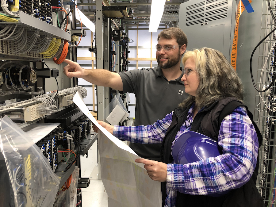 Josh Mabry points towards electrical equipment while standing next to Noelle Currey who is holding a hard hat and a large sheet of paper.