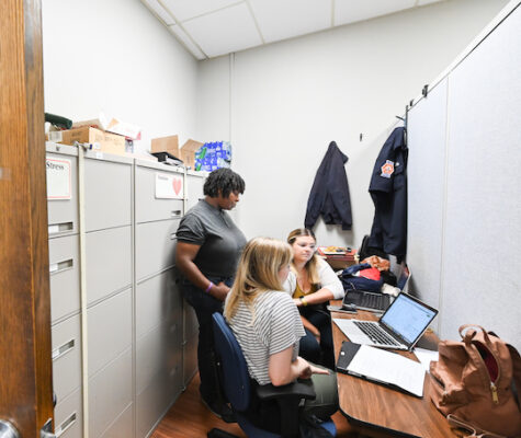 The students sit in an office while the director stands and all three look at their laptop. The door to the office reads, "Chattanooga Fire Department."