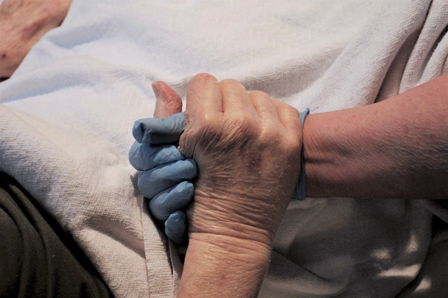 Ramona Grove holds her father’s hand as he lay in bed. She wore gloves as instructed by the care facility in response to COVID-19.
