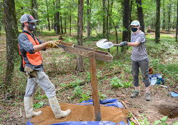 Assistant Professor Morgan Smith holds the sifter while senior Savannah Neely shovels dirt into it.