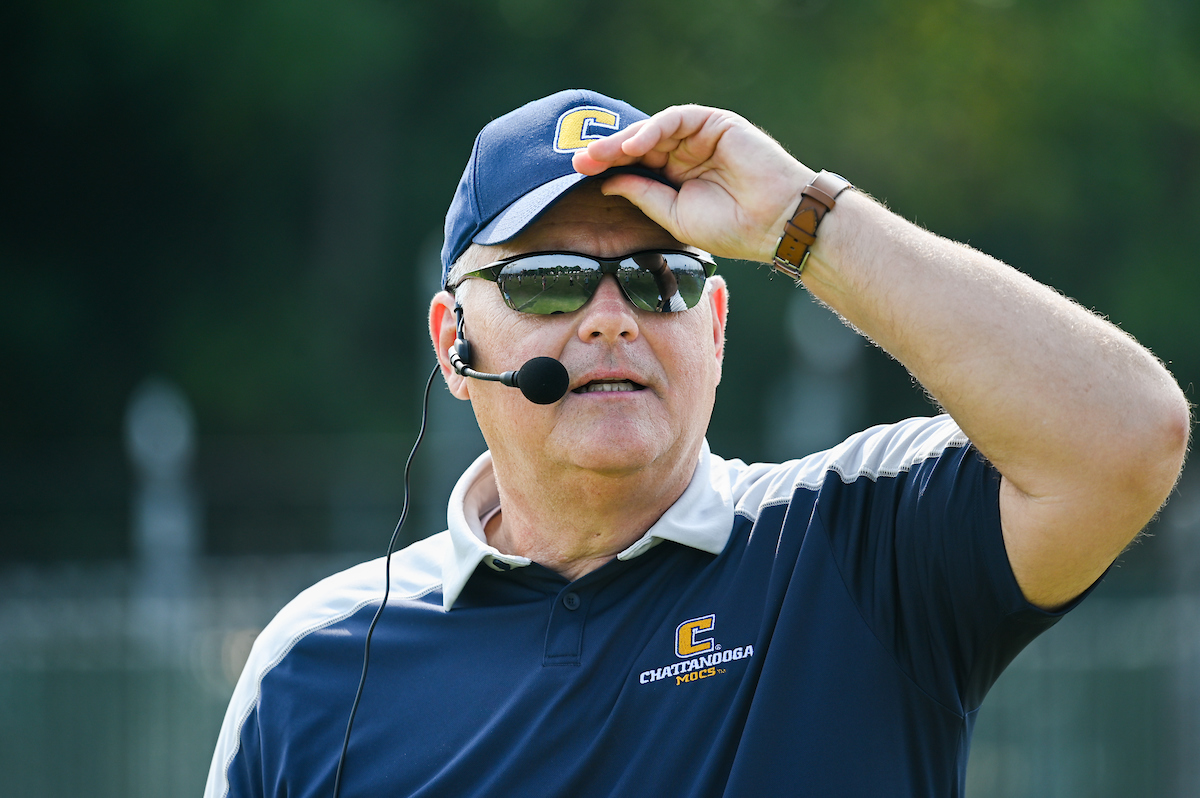 A man wearing sunglasses, headset, and a cap with "C" logo, hands his hand to his cap.