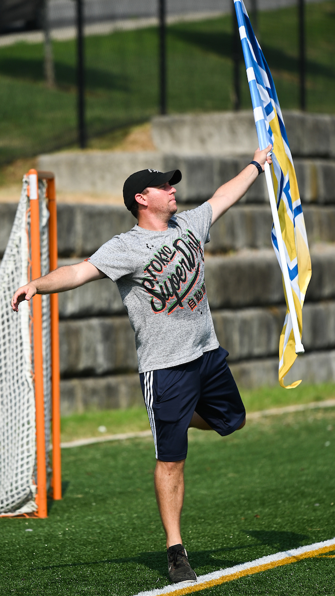 Marching Mocs member waves a blue and yellow flag on a sports field. A goal net and stone wall are in the background.