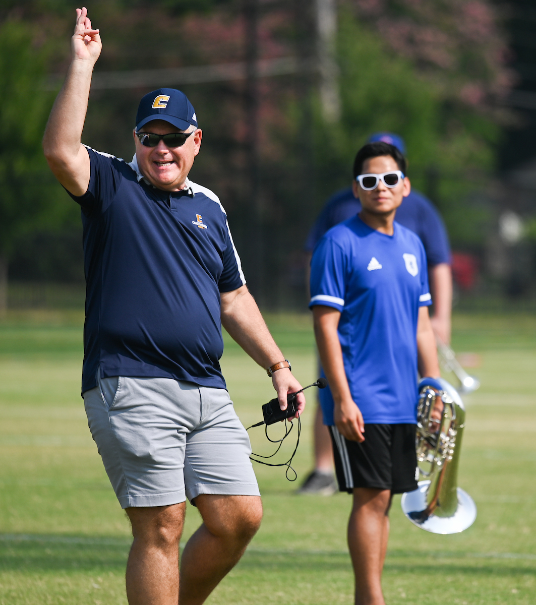 Man in a blue cap and shorts gestures with one hand up. A young man holding a brass instrument stands beside him.