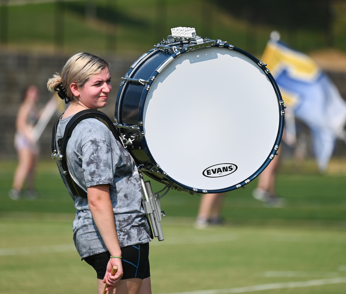 A member of Marching Mocs stands on a grassy field carrying a large bass drum.