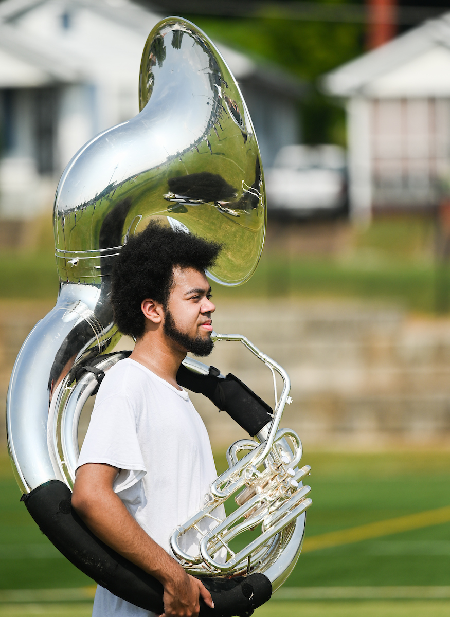 Marching Mocs member carries a sousaphone over his shoulder, outside on a grassy field.