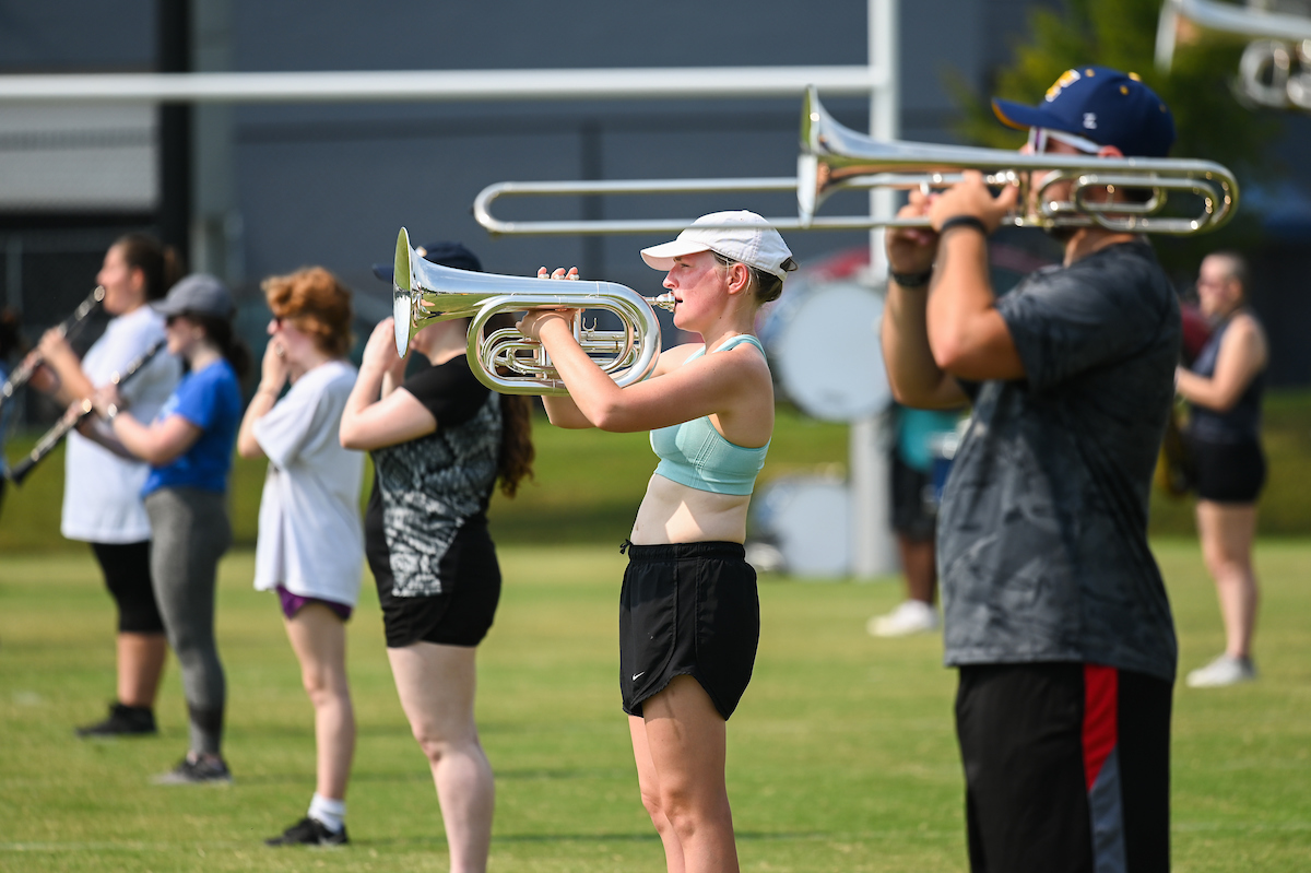 Marching Mocs members play brass and woodwind instruments outside, lined up on a grassy field.