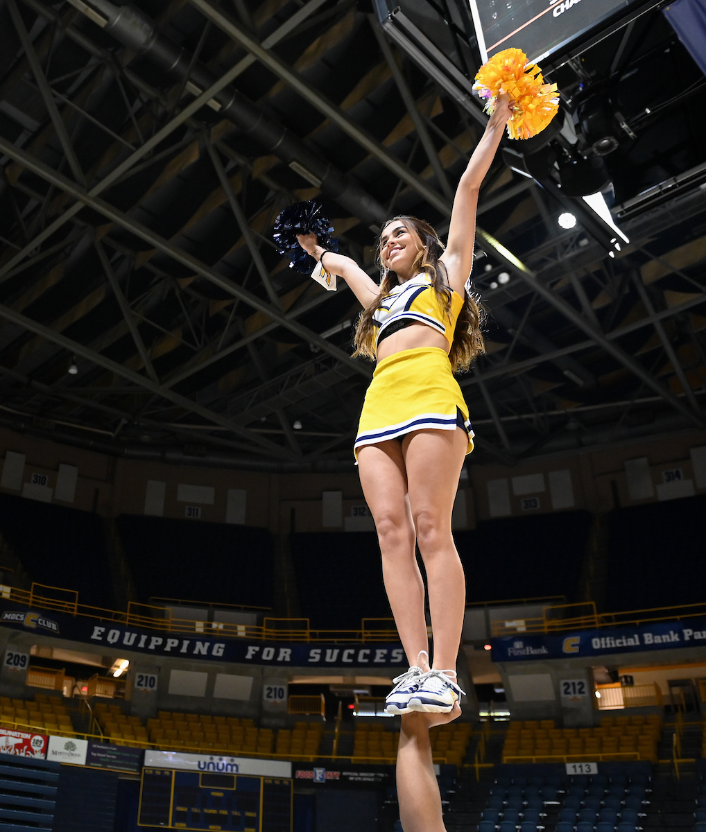 A cheerleader stands poised atop a partner's hand, holding pom-poms above her head.