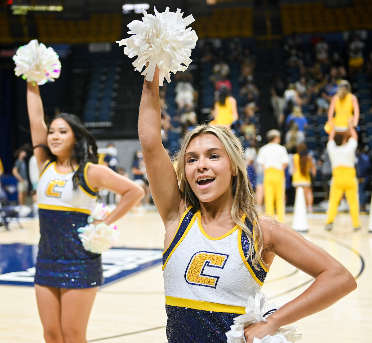 Cheerleaders holding pom-poms smile with arms raised on the basketball court.