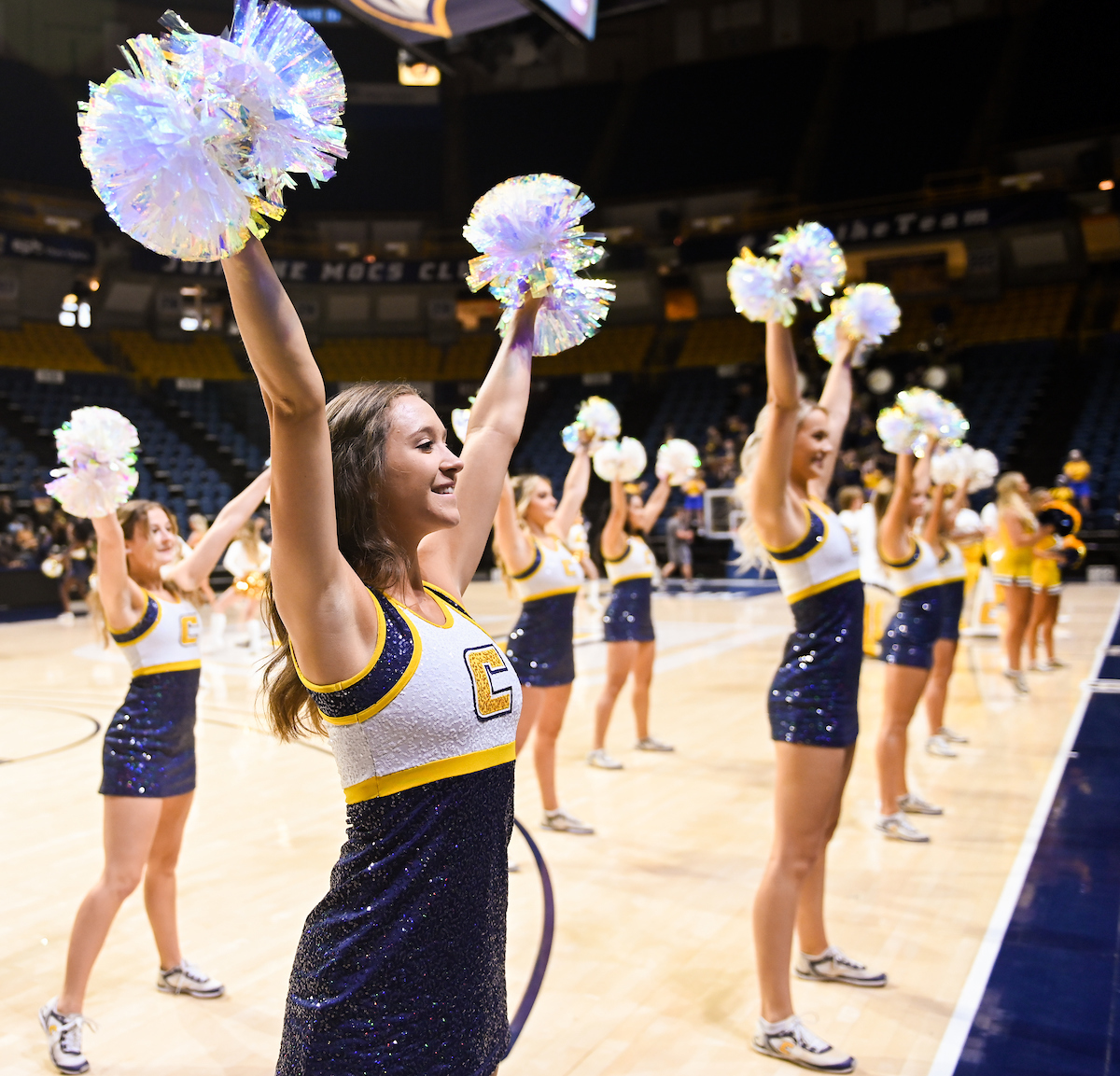Cheerleaders holding pom-poms smile with arms raised on the basketball court.