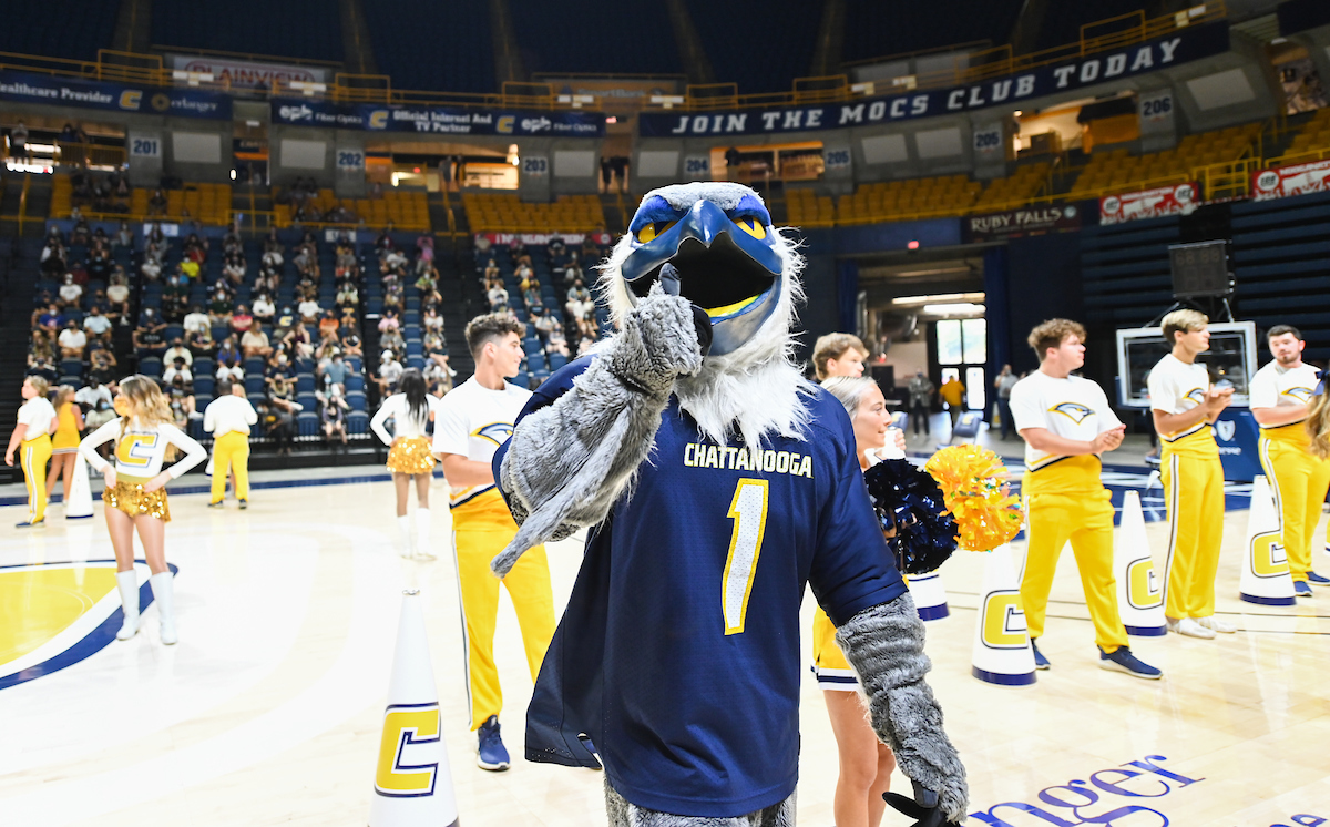 Scrappy posing with a thumbs-up on the McKenzie Arena floor. Cheerleaders and students are seen in the background.