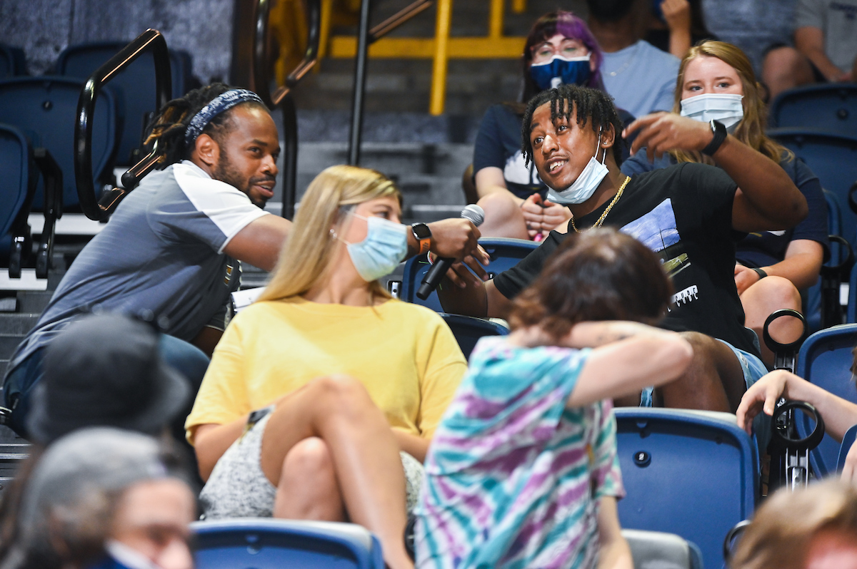 An audience in McKenzie Stadium, with a person holding a microphone interviewing a student.