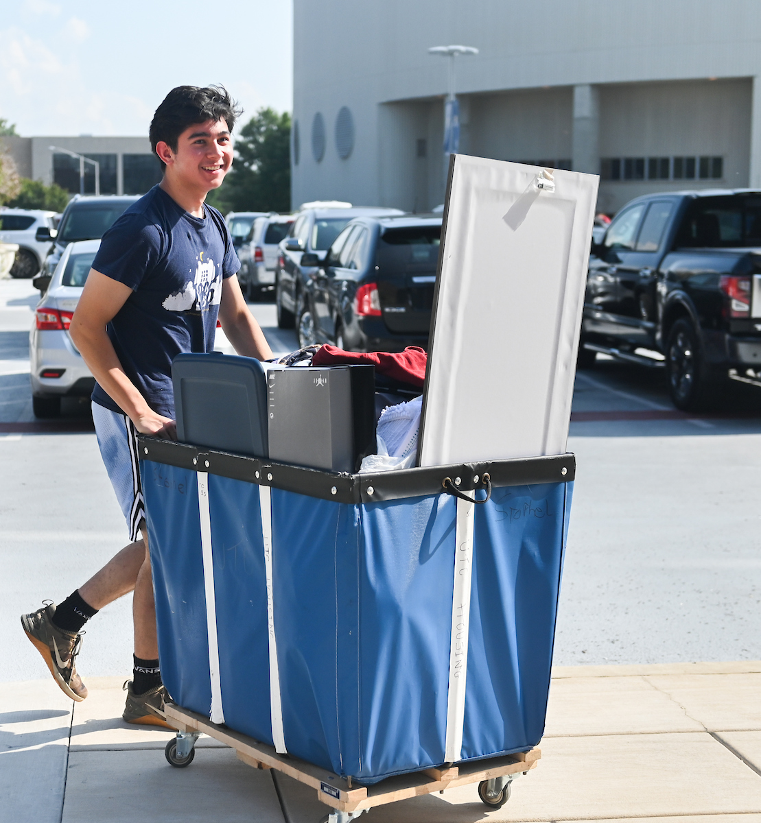 Student smiles while pushing a large blue cart filled with belongings in a parking lot during Operation Move-In.