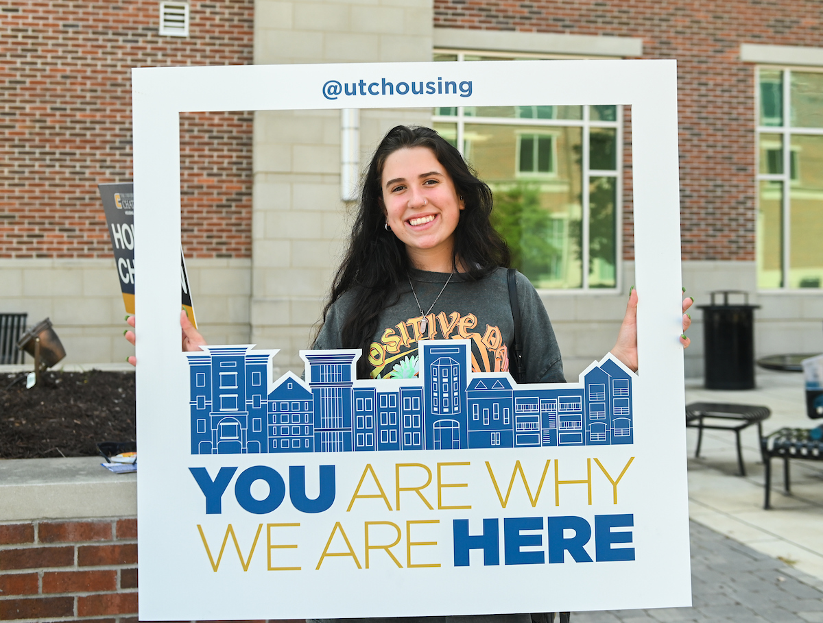 Student holds a frame with the text "@utchousing" and "YOU ARE WHY WE ARE HERE" in blue and gold, standing in front of a brick building.