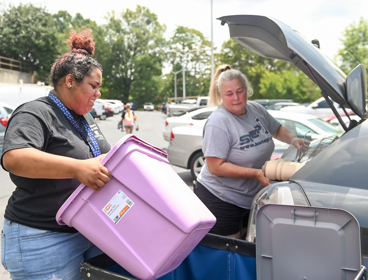 Two women unload a car trunk with a purple storage bin in a parking lot.
