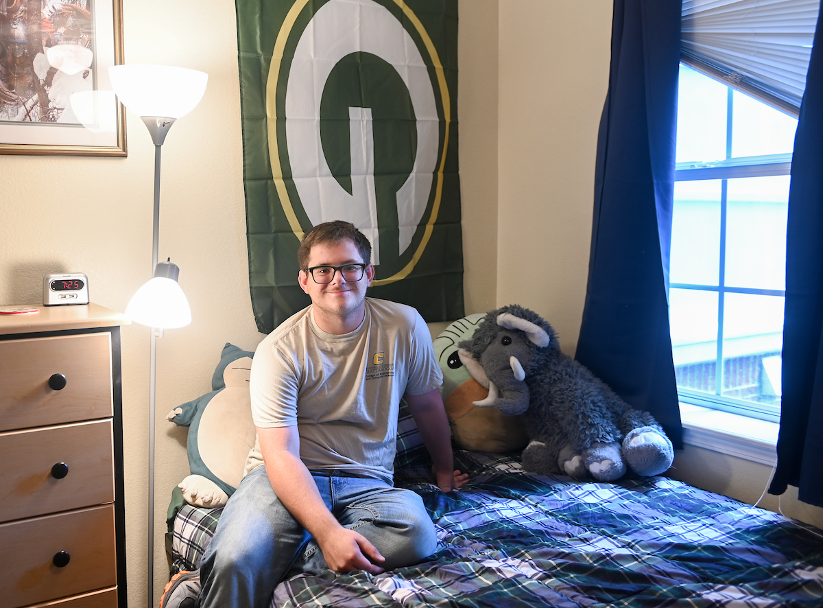 A student sits on a bed with a Green Bay Packers flag on the wall and plush toys beside them.