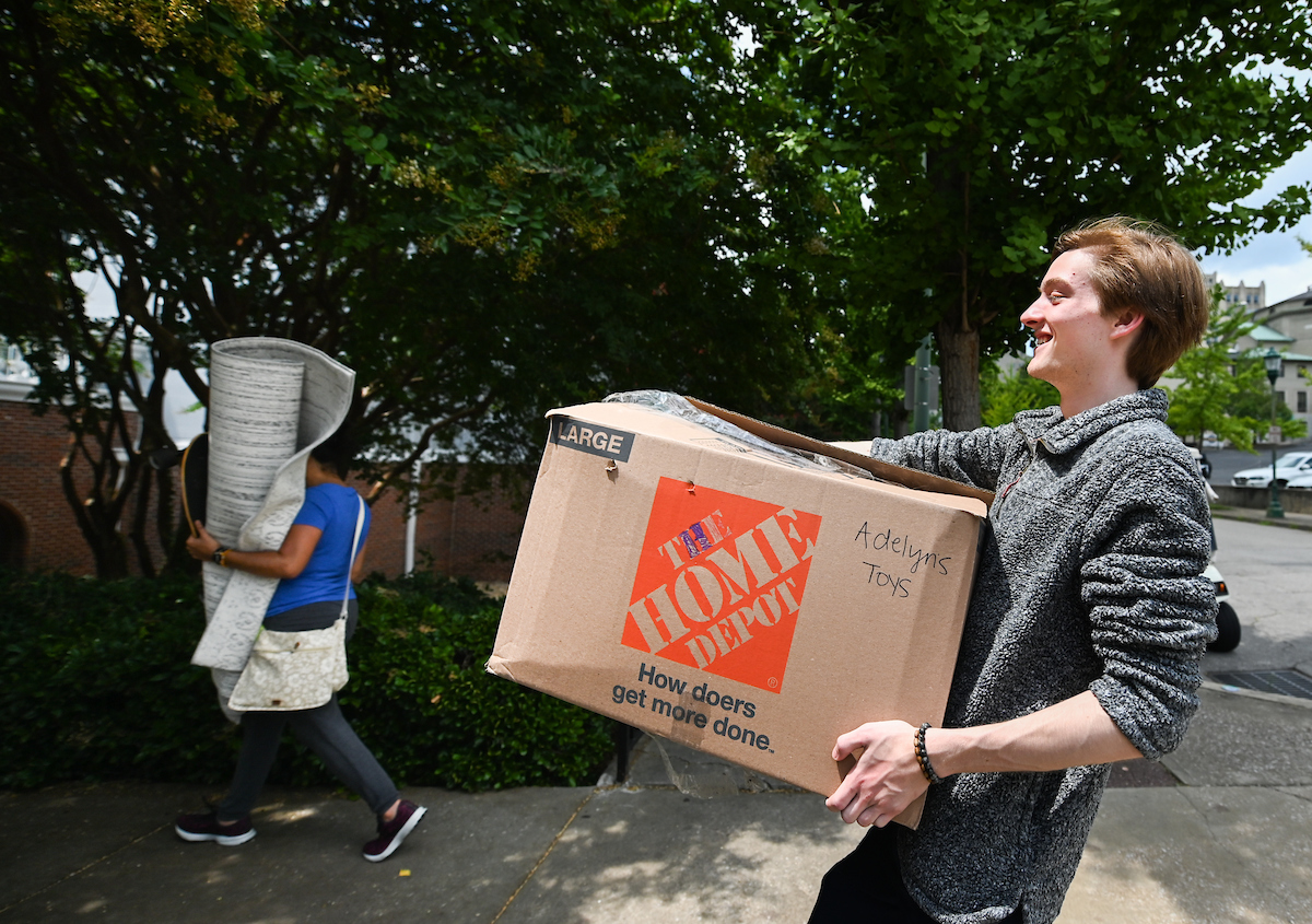 Student carries a large Home Depot box labeled "Adelyns Toys" smiling as he walks down a sidewalk.