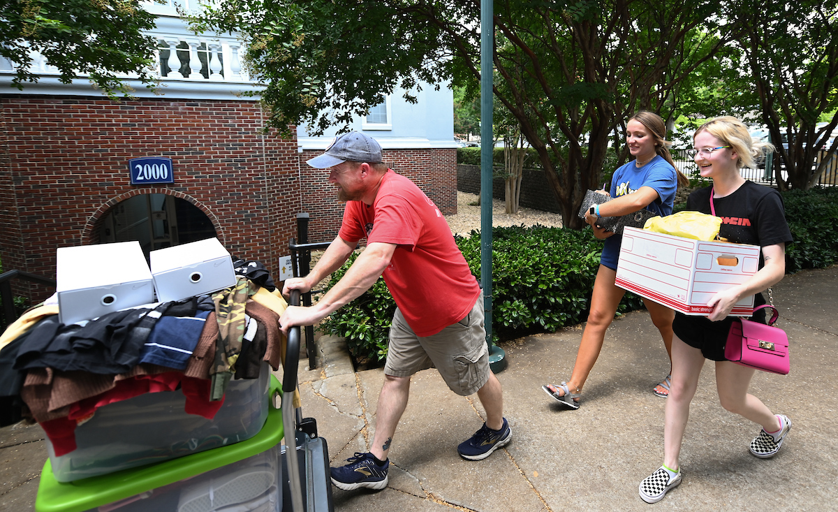 A man pushes cart loaded with boxes and clothes. Two women follow, carrying boxes.