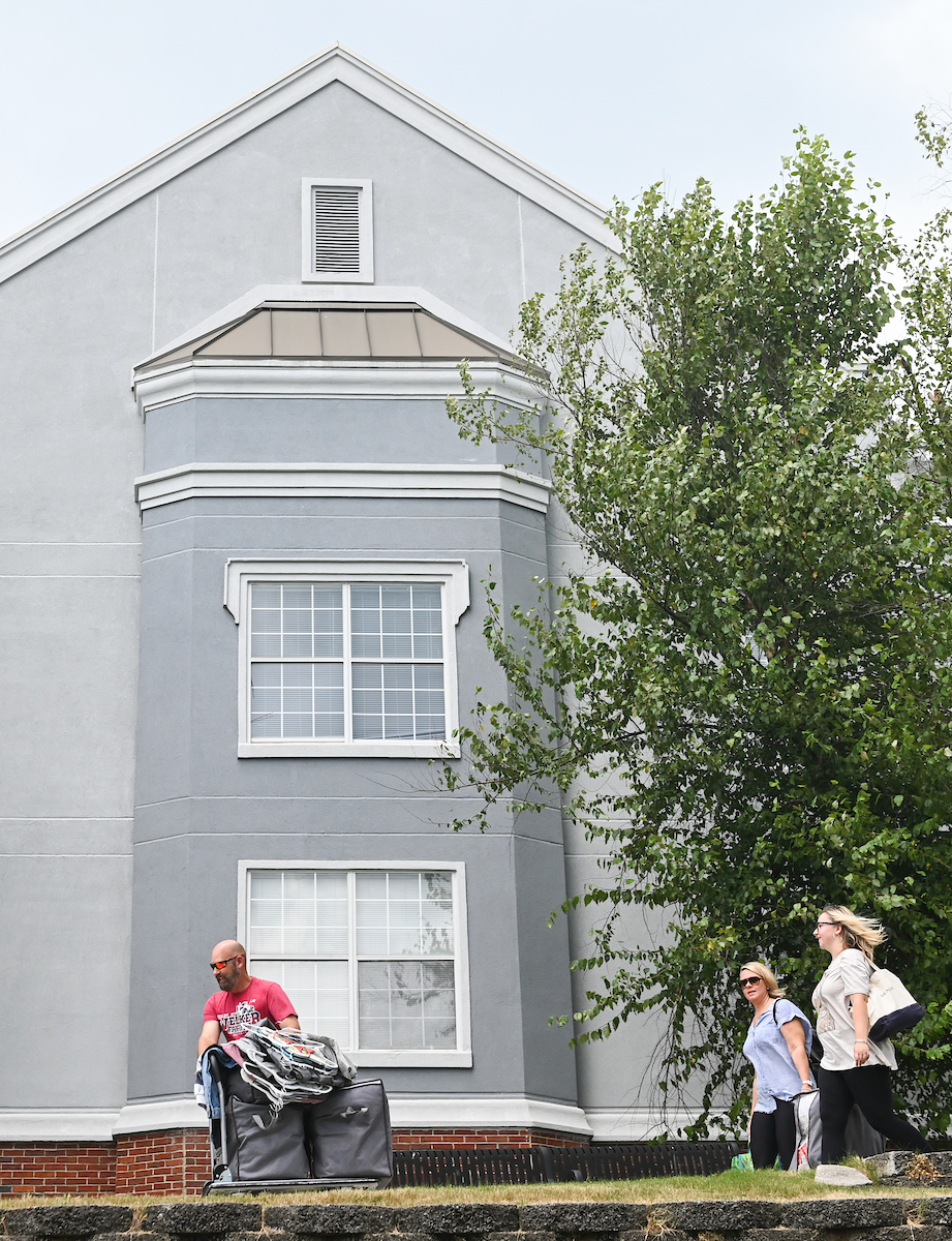 A man pushes a dolly with items in front of a gray building. Two women walk alongside.