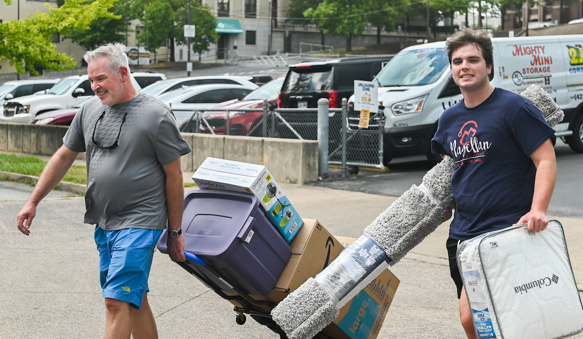 Family moves items with a dolly in a parking lot, carrying boxes and a rug.