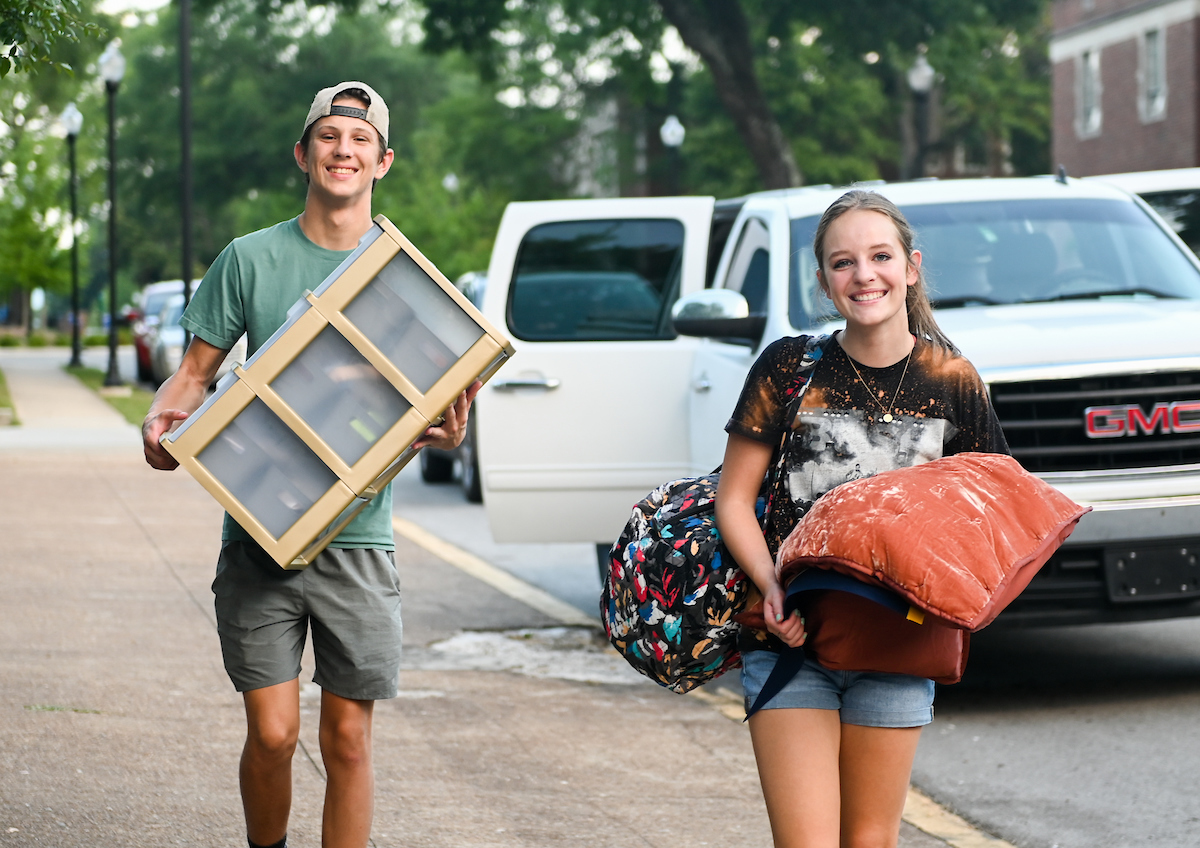 Students smiling while carrying belongings along a sidewalk. Parked cars are behind them.