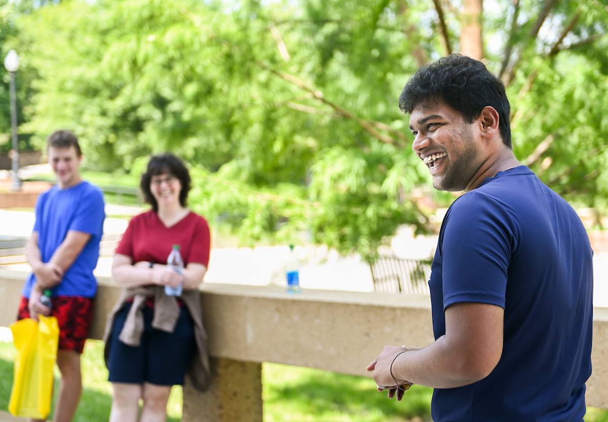 A man smiles while looking to the side, with two people in the background leaning on a concrete railing.