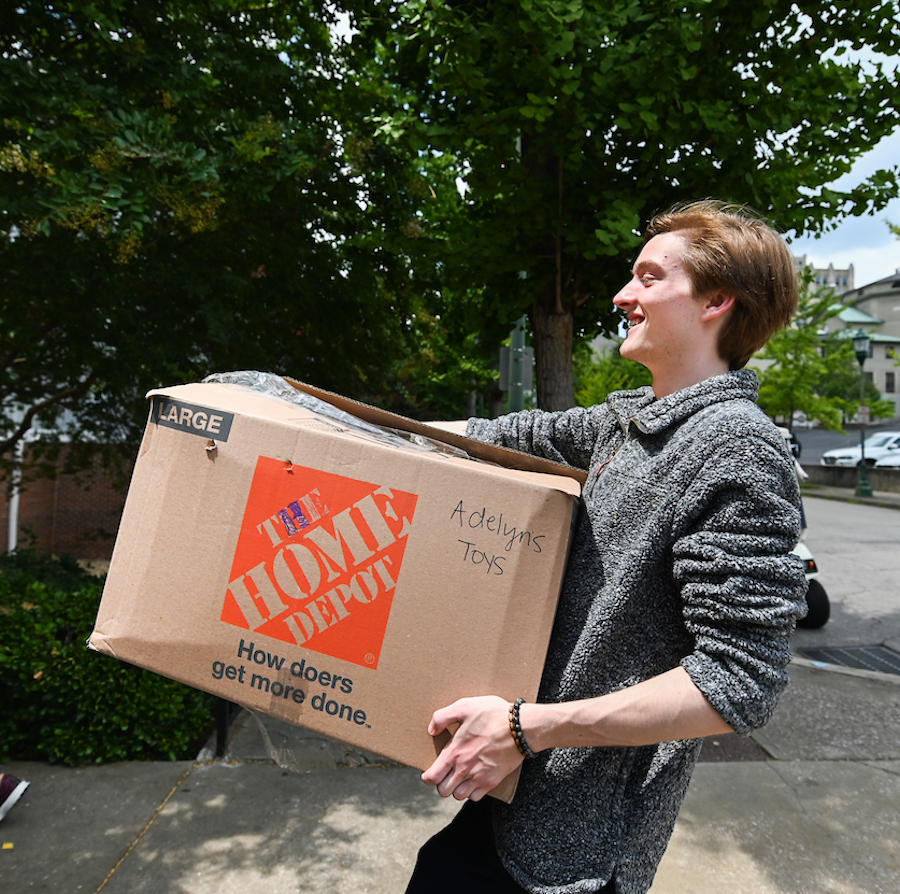Student carries a large Home Depot box labeled "Adelyns Toys" smiling as he walks down a sidewalk.