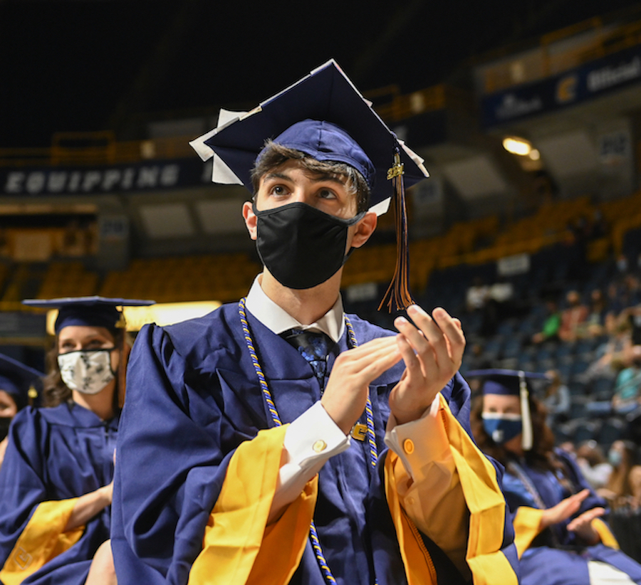 Graduates in blue caps, gowns and face masks sit in rows at graduation ceremony, clapping.