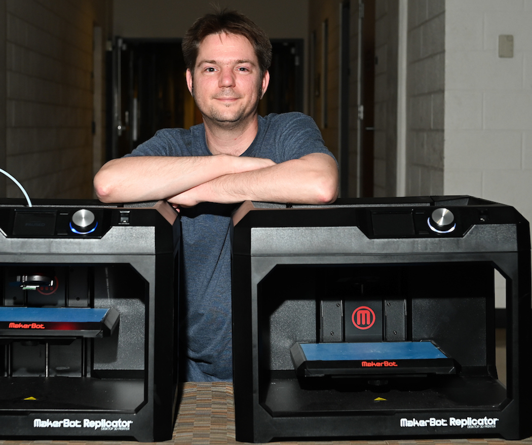 Assistant Professor Trevor Elliot stands smiling behind two MakerBot Replicator 3D printers.