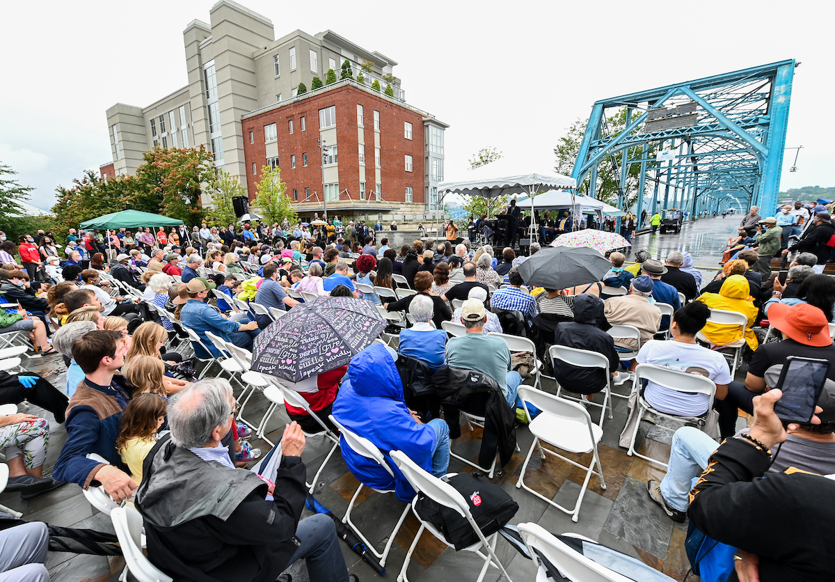 A large crowd of people, some holding umbrellas, sitting outdoors near a bridge and tall buildings.