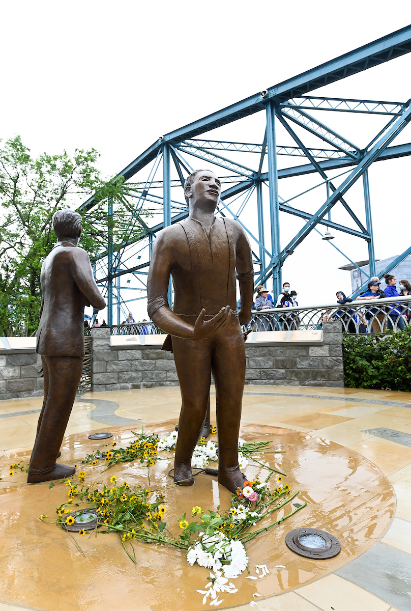 Bronze statues of two standing figures on a wet pedestal, adorned with yellow and white flowers.